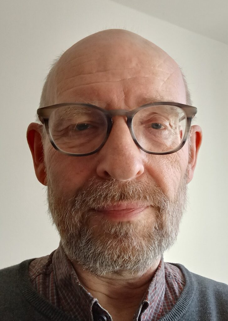 Ein Mann mittleren Alters mit Bart und Brille, der ein kariertes Hemd und einen grauen Pullover trägt, blickt vor einem schlichten hellen Hintergrund in die Kamera.