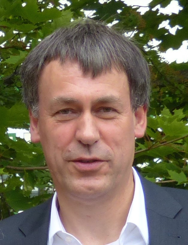 Ein Mann mittleren Alters mit kurzen braunen Haaren, der eine dunkle Jacke und ein weißes Hemd trägt, steht im Freien vor grünen, belaubten Bäumen.