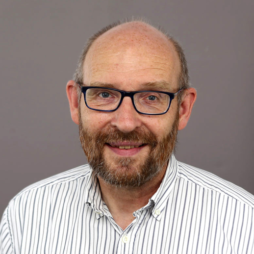 Ein Mann mittleren Alters mit einer Glatze, einer Brille und einem Bart lächelt in die Kamera. Er trägt ein weißes Hemd mit dünnen schwarzen Streifen und steht vor einem schlichten grauen Hintergrund.