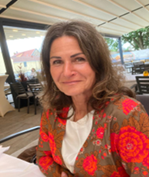 Eine Frau mit gewelltem braunem Haar, die eine braune Jacke mit roten und rosa Blumenmustern trägt, sitzt an einem Tisch in einem Restaurant im Freien mit einem Vordach und unscharfem Hintergrund.