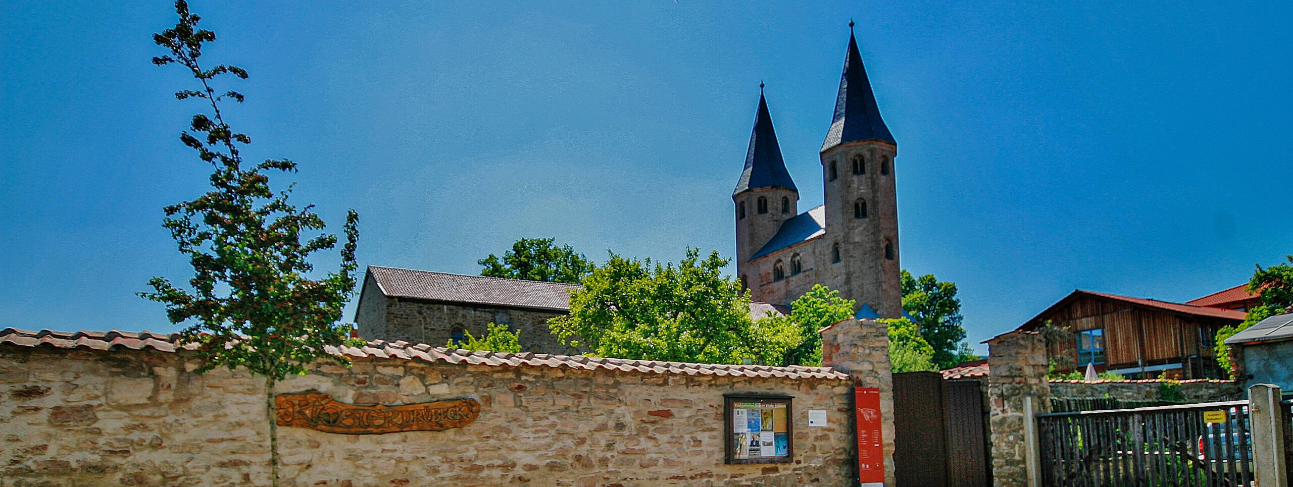 Eine Steinmauer mit einem hölzernen Schild und einer Hinweistafel verläuft vor einer historischen Kirche mit zwei hohen Türmen, teilweise umgeben von Bäumen und rustikalen Gebäuden unter einem klaren blauen Himmel.