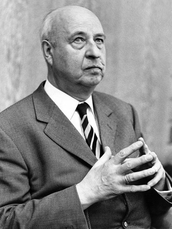 Ein älterer Mann im Anzug und mit gestreifter Krawatte sitzt mit verschränkten Fingern da und blickt nachdenklich nach oben. Das Foto ist in schwarz-weiß gehalten.