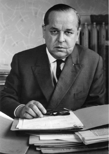 Ein Mann im Anzug sitzt an einem Schreibtisch mit Dokumenten und Brille in der Hand und schaut ernst in die Kamera. Im Hintergrund ist eine Büroumgebung zu sehen.